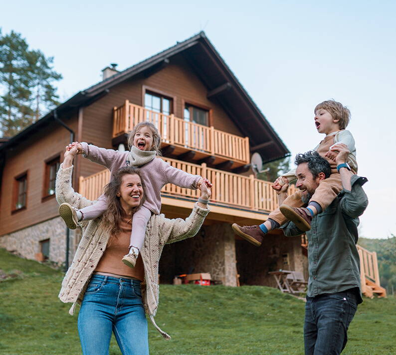 Familie vor eigenem Haus in naturnaher Umgebung Eine Familie steht auf einer grünen Wiese vor einem modernen Holzhaus mit Balkon und Steinsockel. Im Hintergrund sind Bäume und blauer Himmel sichtbar, was eine idyllische Wohnsituation darstellt.