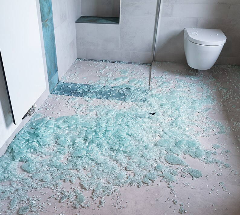 Shards from a shattered glass shower wall cover the floor of a modern bathroom. The fragments reflect the light, making the damage clearly visible.
