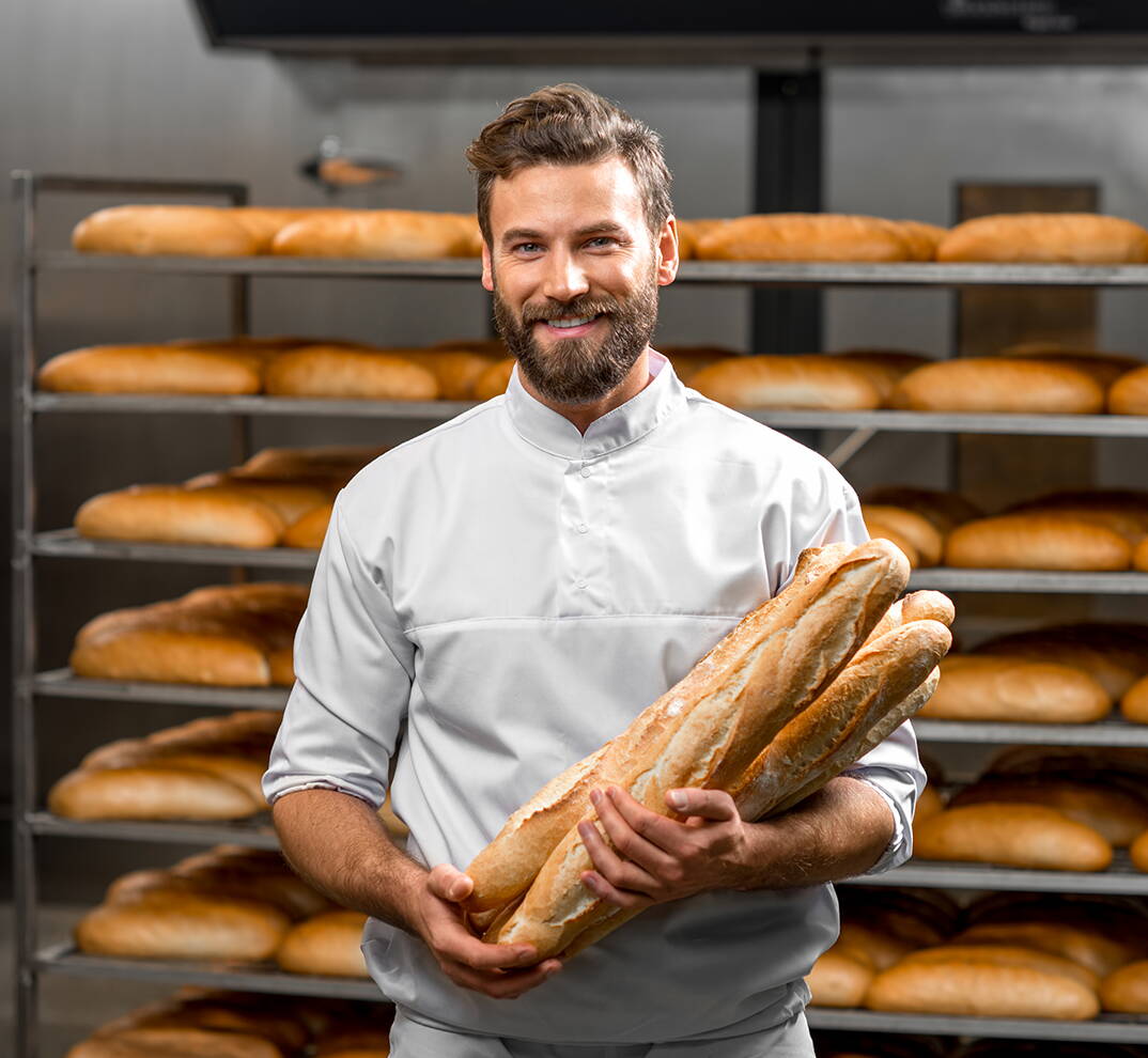 Eine Person hält mehrere frisch gebackene Baguettes vor einem Regal voller Brote.